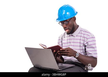 young man engineer wearing hard hat sitting on chair and checking notes in notebook using laptop. Stock Photo