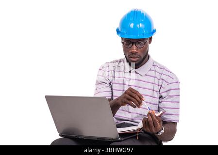 young man engineer wearing safety helmet sitting on chair and working with laptop and notebook in hand. Stock Photo