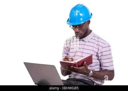young man engineer wearing hard hat sitting on chair and taking notes in notebook using laptop. Stock Photo