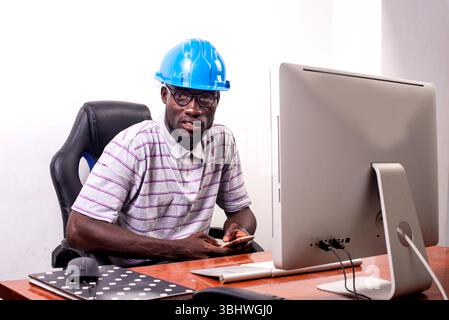 young man engineer wearing blue safety helmet sitting at desk sitting at desk using mobile phone. Stock Photo