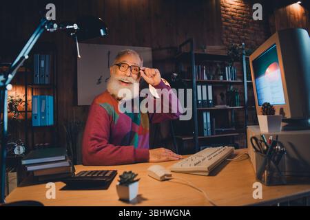Elderly man working with a retro computer in a cozy home office showing cheerful expression under warm evening light Stock Photo