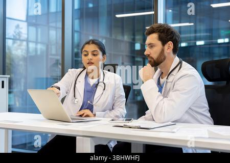 Two doctors, one female and one male, are looking at a laptop screen in a modern office setting, collaborating on a case. Stock Photo