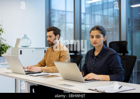 Two colleagues work at a desk using laptops in a modern office setting. Stock Photo