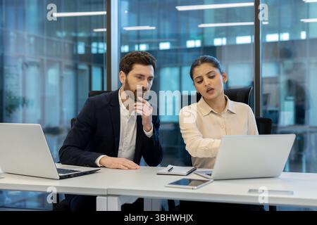 A man and a woman are collaborating on a laptop, discussing a project in a modern office setting. Stock Photo