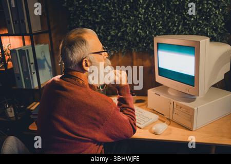 Senior man working thoughtfully on a vintage computer in a cozy home office during the evening Stock Photo