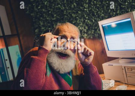 Elderly man in sweater smiling and working at computer in cozy home office setting with focus on the retro screen Stock Photo