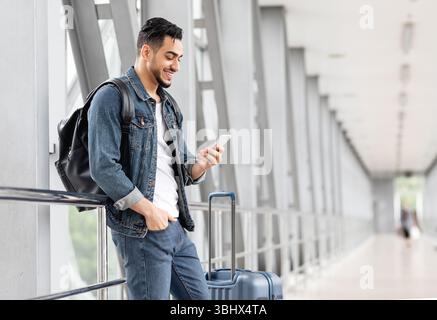 Smiling Arab Male Traveller With Smartphone And Luggage Waiting In Airport Stock Photo