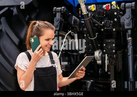 Female technician working on digital tablet while talking on phone near tractor Stock Photo