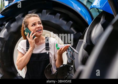 Female agricultural worker looking puzzled while talking on phone next to tractor Stock Photo