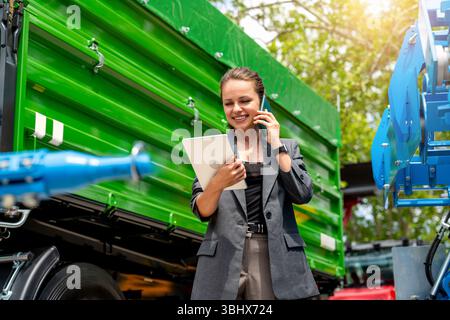 Businesswoman in suit communicating with client on phone while holding digital tablet at farm equipment dealer Stock Photo