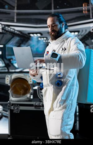 Astronaut researcher verifying mission parameters using laptop, carrying out research aboard. Scientist wearing cosmonaut gear in spaceship updating flight log using notebook device Stock Photo