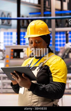 Automation female engineer in high tech industrial facility, ensuring efficiency of solar panels and machinery. African american woman optimizing system performance in smart manufacturing plant. Stock Photo