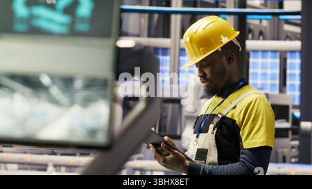 Engineer oversees solar panel production lines equipment using tablet app. Worker maintains photovoltaic assembly systems in automated industry 4.0 factory using digital device application, camera B Stock Photo