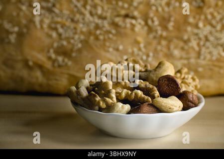Walnuts, cashews, hazelnuts, and other mixed nuts filling a small white bowl, with a loaf of bread blurred in the background Stock Photo