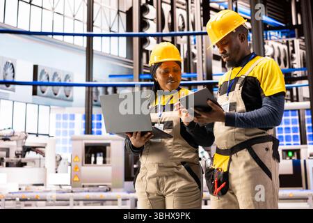 Engineers using digital interfaces on laptop and tablet to monitor and enhance efficiency in automated factory. Industry 4.0 innovation powers industrial automation in smart manufacturing facility. Stock Photo