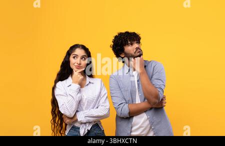 Portrait of pensive indian man and woman thinking and looking up at free space, touching chin, yellow background Stock Photo
