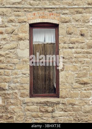 Building built 1900 with sandstone block walls, Windows have red brick keystone & are covered with old drapes. Curtains are veiled on glass window. Stock Photo