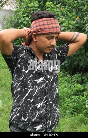 Young South Asian laborer with a traditional red bandana (gamcha) tied on his forehead, posing with hand on his head. Concept of hard work and rural l Stock Photo