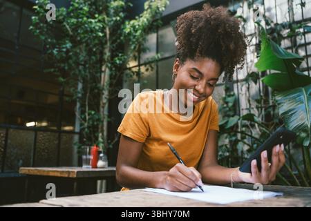 Smiling woman using smartphone and writing notes at table in cafe with plants Stock Photo
