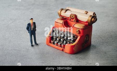 Miniature Businessman Standing Next to Oversized Vintage Typewriter on Gray Surface Stock Photo