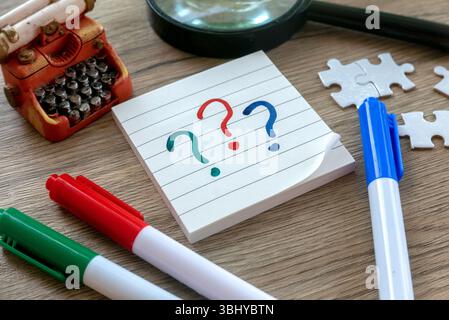 Colorful Question Marks on Notepad with Pens, Puzzle Pieces, Magnifying Glass, and Mini Typewriter on Wooden Desk Stock Photo