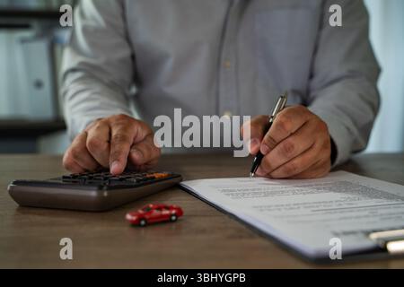 An insurance on paperwork assurance person gray shirt is working desk, using calculator and signing document with pen, with small red toy car nearby Stock Photo