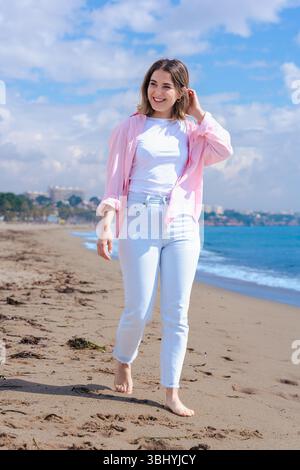 Smiling Woman Waving Hand at Camera While Standing on Sunny Beach Near Ocean Stock Photo