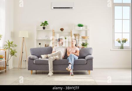 Happy couple relaxing on sofa in living room, talking and enjoying cool air from air conditioner. Stock Photo