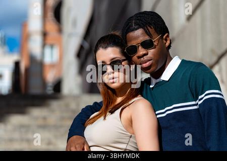 Interracial couple wearing stylish sunglasses, posing together on city steps, exuding love and happiness in a trendy urban setting Stock Photo