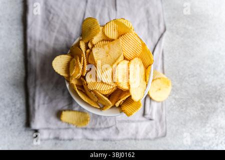 A bowl filled with delicious homemade potato chips, showcasing a golden, crispy texture, resting on a soft gray cloth, perfect for a snack or a casual Stock Photo