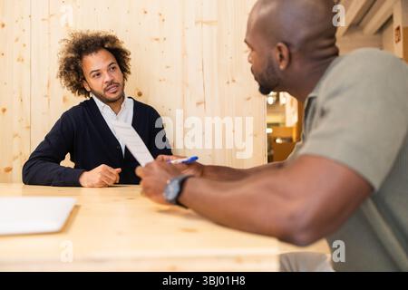 Two coworkers engage in a focused discussion in a modern coworking space. The environment features natural wood decor, fostering creativity and collab Stock Photo