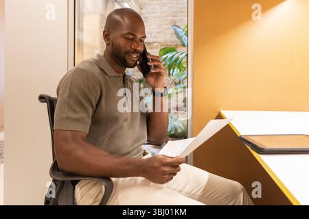 Professional man in a modern coworking space in Barcelona, engaged in a business call while reviewing documents. The setting is vibrant, creative, and Stock Photo