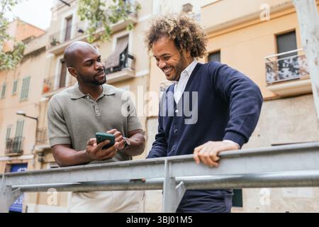 Two men enjoying a break outside a coworking space share a smartphone, engaged in a friendly and relaxed conversation, surrounded by an urban setting Stock Photo