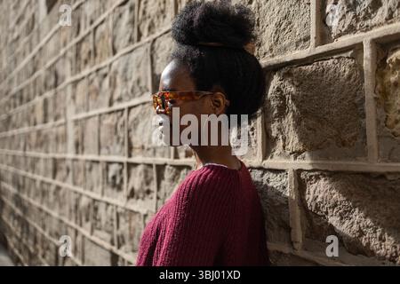 A stylish woman in a burgundy sweater wearing sunglasses stands confidently against a textured stone wall, casting a shadow in the warm sunlight of a Stock Photo