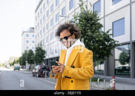 A fashionable man with an afro hairstyle and sunglasses, wearing a stylish yellow coat, smiling while using his smartphone outdoors in an urban settin Stock Photo