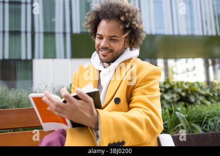A cheerful Afro man with curly hair, wearing a bright yellow coat, enjoying a peaceful moment reading a book on a bench in an urban outdoor setting. Stock Photo