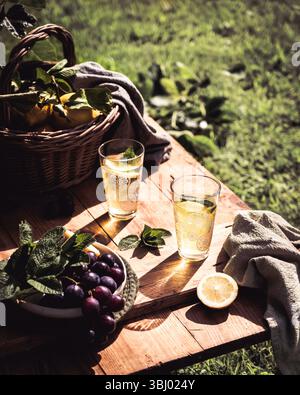 Two glasses of homemade lemonade sit on a rustic wooden table surrounded by fresh grapes and a wicker basket of lemons, with a garden providing a vibr Stock Photo