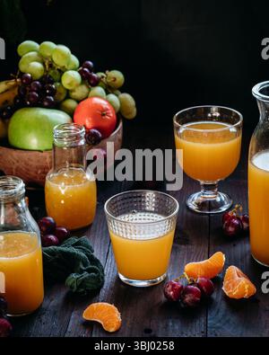 Assorted glasses and bottles of fresh orange juice surrounded by vibrant fruits, including grapes and oranges, on a rustic wooden table, creating a re Stock Photo
