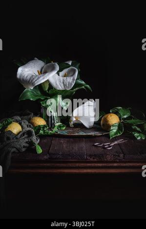 Elegant calla lilies in a glass vase complement ripe lemons on a rustic wooden table. The dark background accentuates the textures and contrasts of th Stock Photo