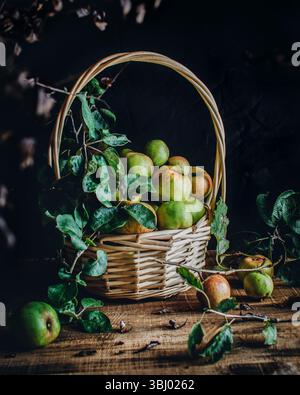 A rustic wicker basket filled with fresh green apples sits on a wooden table. Surrounded by leaves and twigs, the scene evokes a cozy, autumn harvest Stock Photo