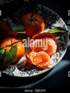 A group of ripe tangerines with vibrant green leaves sits elegantly on a decorative plate. One tangerine is peeled open, revealing its juicy segments Stock Photo