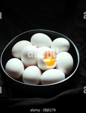 A collection of white eggs sits encircled in a dark bowl on a black background, with one egg cracked open to reveal its bright yellow yolk, adding a p Stock Photo