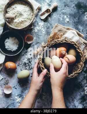 Rustic kitchen scene with hands holding a basket of assorted brown eggs, surrounded by flour, salt, and baking utensils on a textured stone countertop Stock Photo