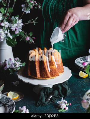 A moist spring lemon bundt cake is elegantly displayed on a cake stand, topped with a drizzle of icing. Surrounded by floral decor, it captures the es Stock Photo