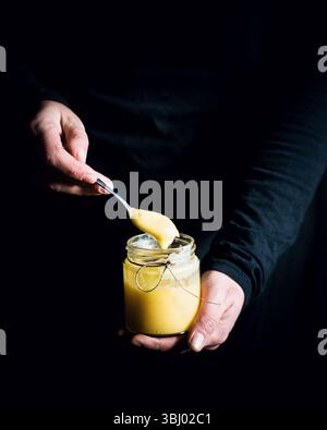 A close up view of homemade lemon curd in a glass jar, with a person holding a spoonful of the creamy, tangy spread. Perfect for baking or as a tasty Stock Photo