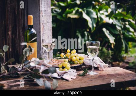 A serene outdoor still life featuring a bottle of white wine, two glasses, and a bunch of fresh grapes on a rustic wooden table, surrounded by lush gr Stock Photo