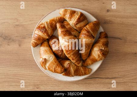 A top view of freshly baked homemade croissants arranged on a white plate, placed on a wooden surface. The golden brown pastries evoke a warm, invitin Stock Photo