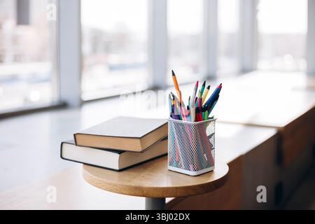 Colorful stationery items such as pencils and pens in a mesh holder alongside two hardcover books on a round wooden table, bathed in natural sunlight Stock Photo