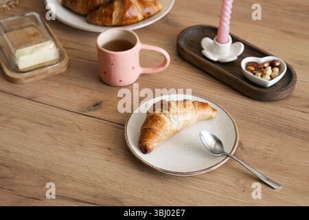 Delicious homemade croissants served on a rustic wooden table with a pink mug filled with coffee, and a dish of assorted nuts for a perfect breakfast Stock Photo