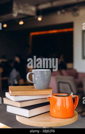 A cozy coffee shop scene with a grey mug placed on a stack of books, accompanied by an orange teapot on a wooden table. The background is softly blurr Stock Photo